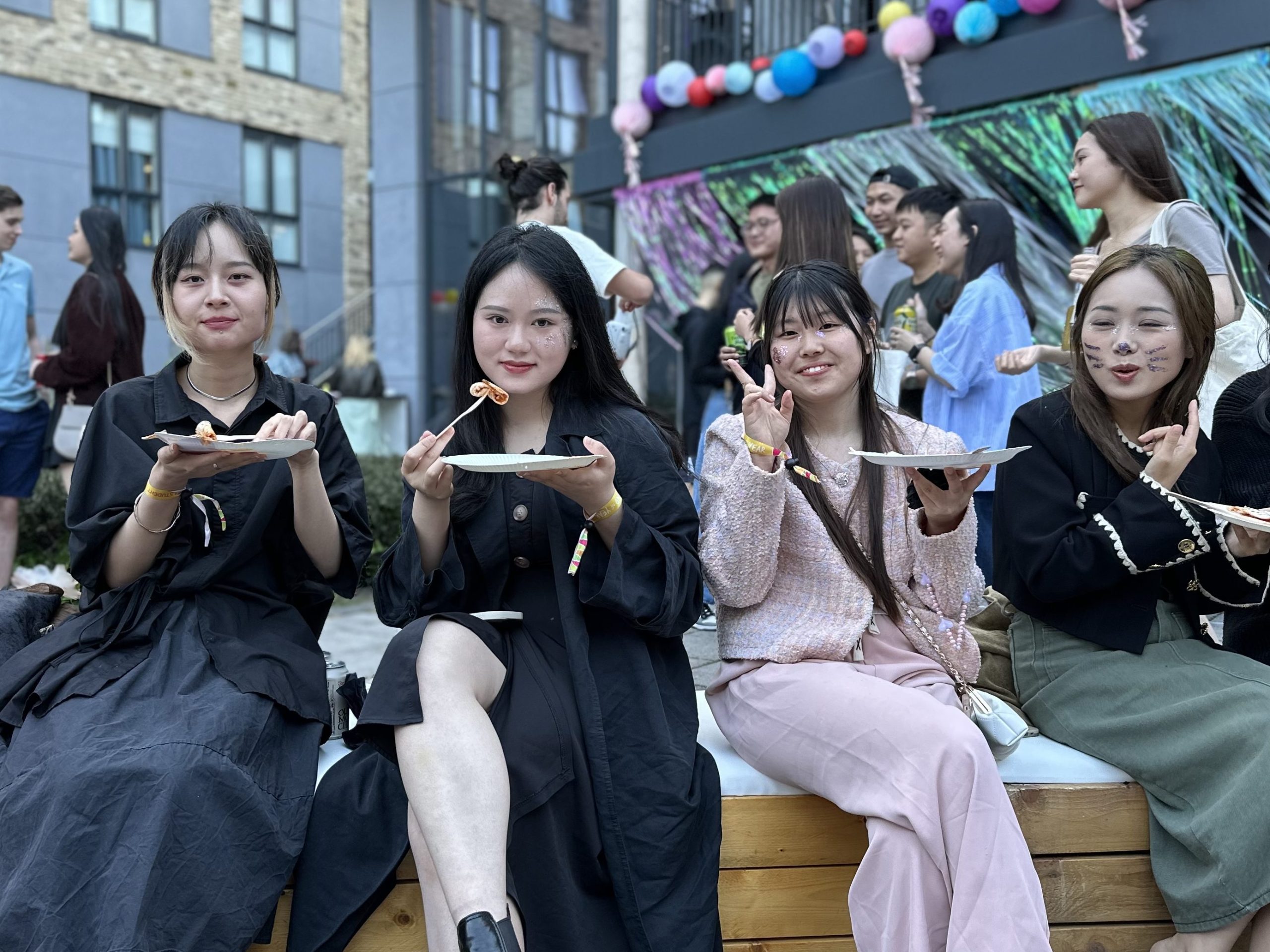 Five women sit on a bench outdoors in Southampton, smiling and eating food from plates, while others mingle by a decorated building—making it a great place to relax after a study session.