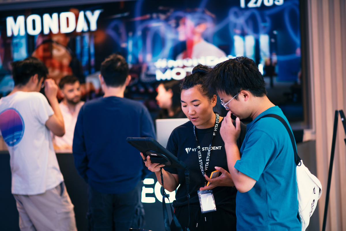 Two people look at a tablet together in the foreground, while three others talk in the background. A large screen behind them displays "MONDAY" in bright letters. The event appears to be related to IE University or student residences.