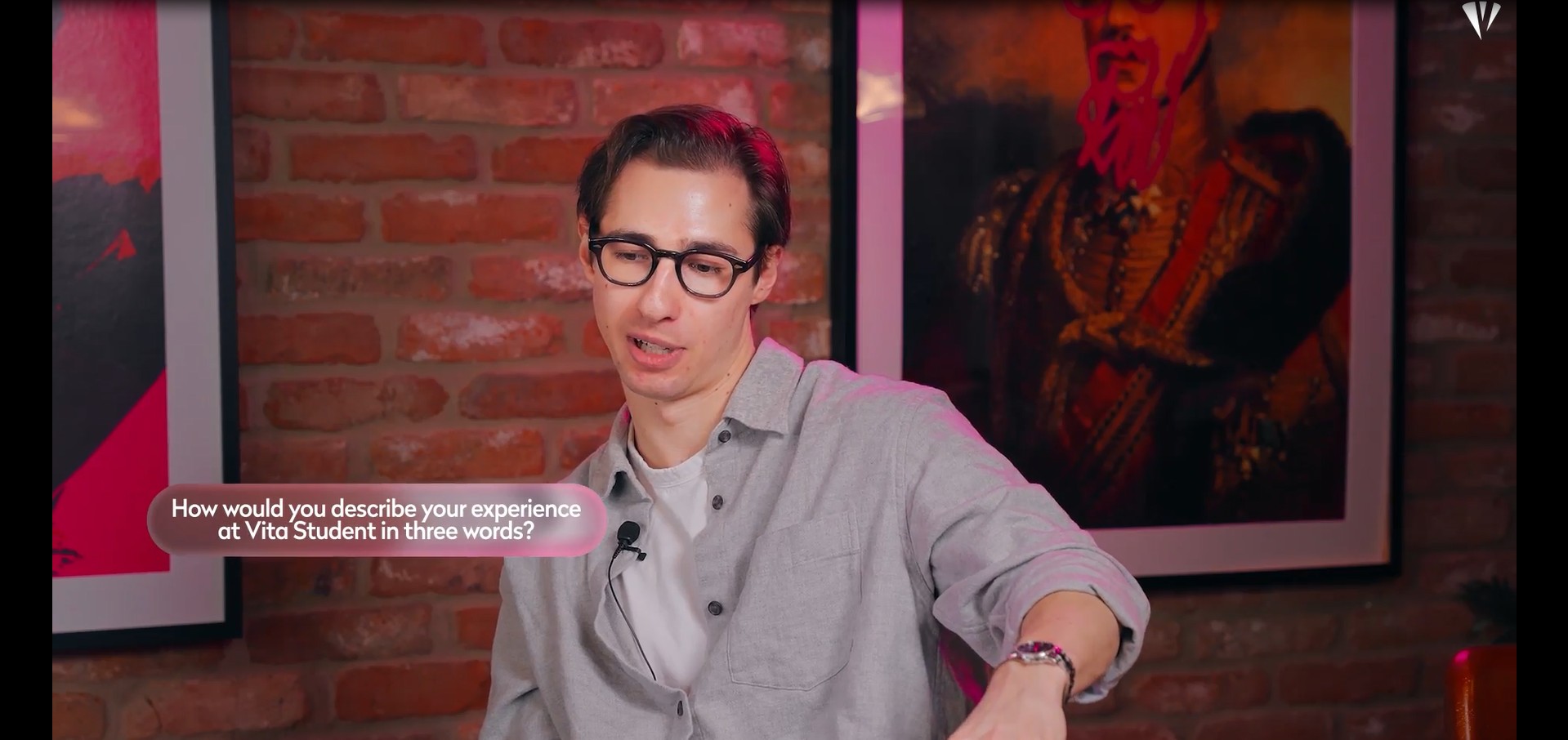 A man wearing glasses and a gray shirt speaks in front of a brick wall with framed art. A text bubble reads, "How would you describe your experience at Vita Student in three words?.