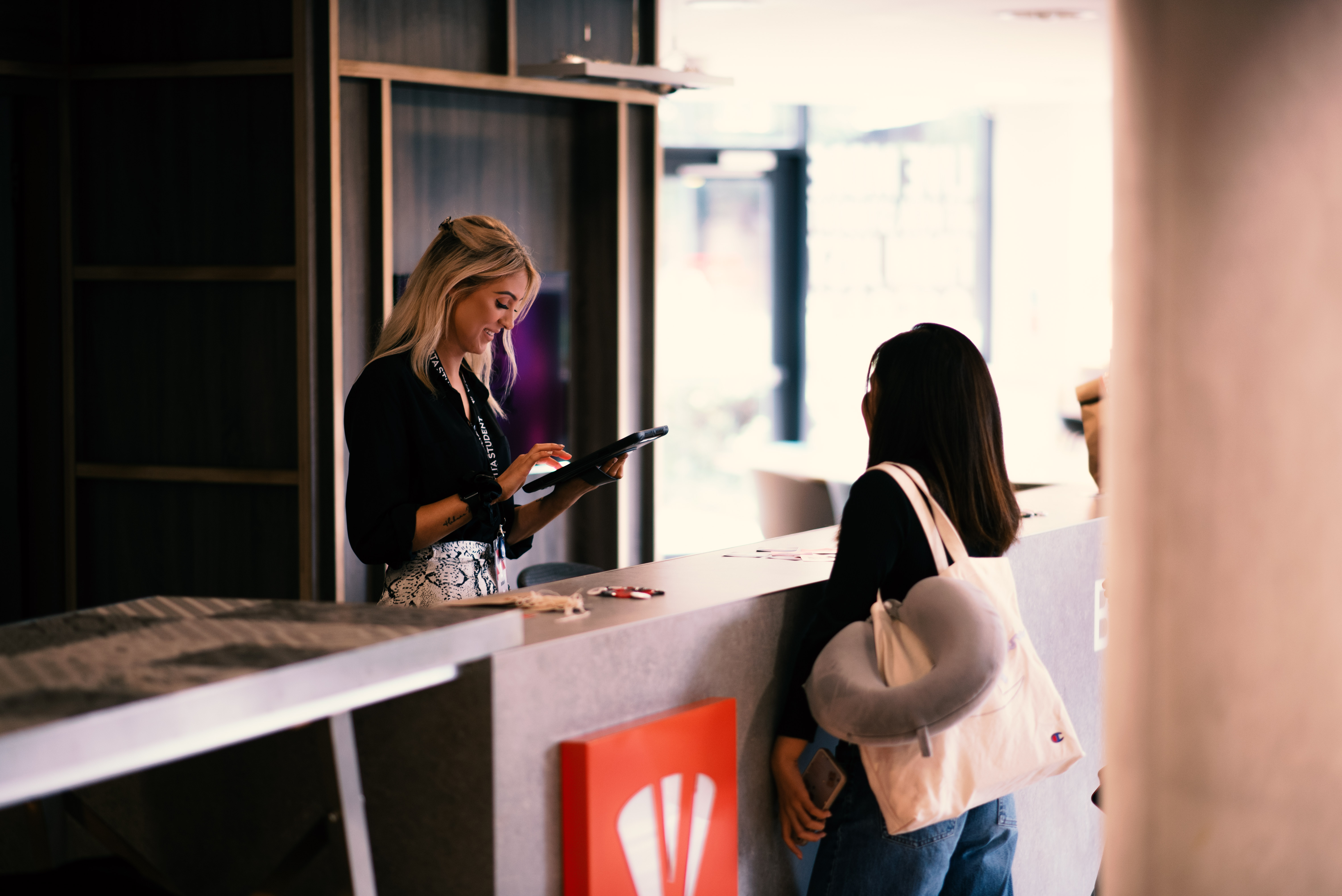 A receptionist stands behind a modern front desk in City Madrid, using a tablet, while a woman with a tote bag and neck pillow over her shoulder stands on the other side, seemingly checking in or asking for information.