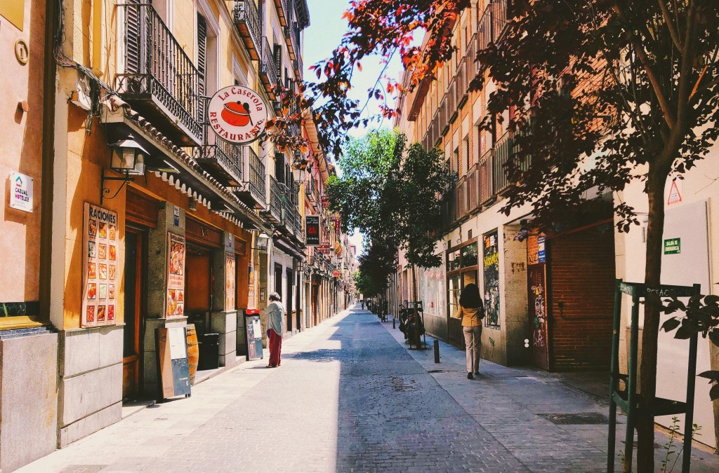 A narrow cobblestone street in a European city, much like those found when exploring where to live in Madrid, is lined with colorful buildings, cafes, and restaurants. Trees provide shade as a few people walk along the quiet, sunlit street.