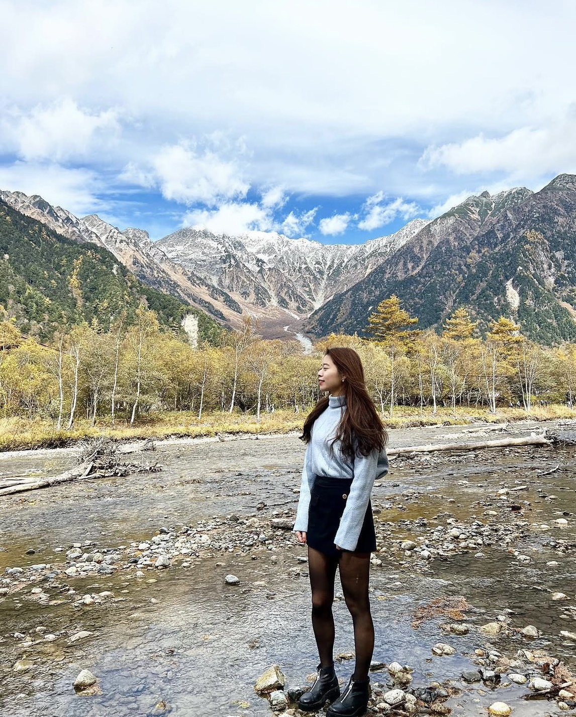 A woman stands on rocky ground by a shallow stream, looking to the side—her pose reminiscent of a Vita Student exploring nature—with mountains, autumn trees, and a partly cloudy sky stretching behind her.