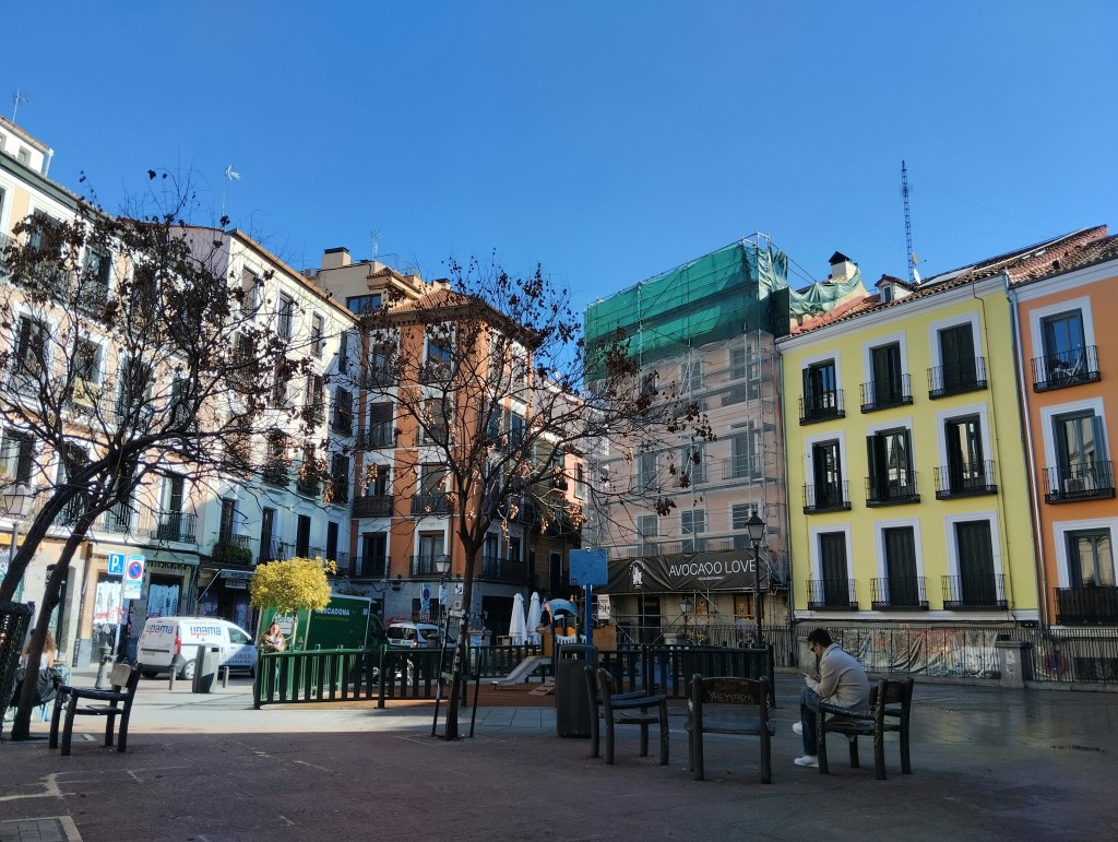 A sunny city square with colorful buildings, leafless trees, benches, and a person sitting alone. One building is covered with scaffolding, and a sign reads "MADRID LOVE." It's a glimpse into the charm of where to live in Madrid under clear blue skies.