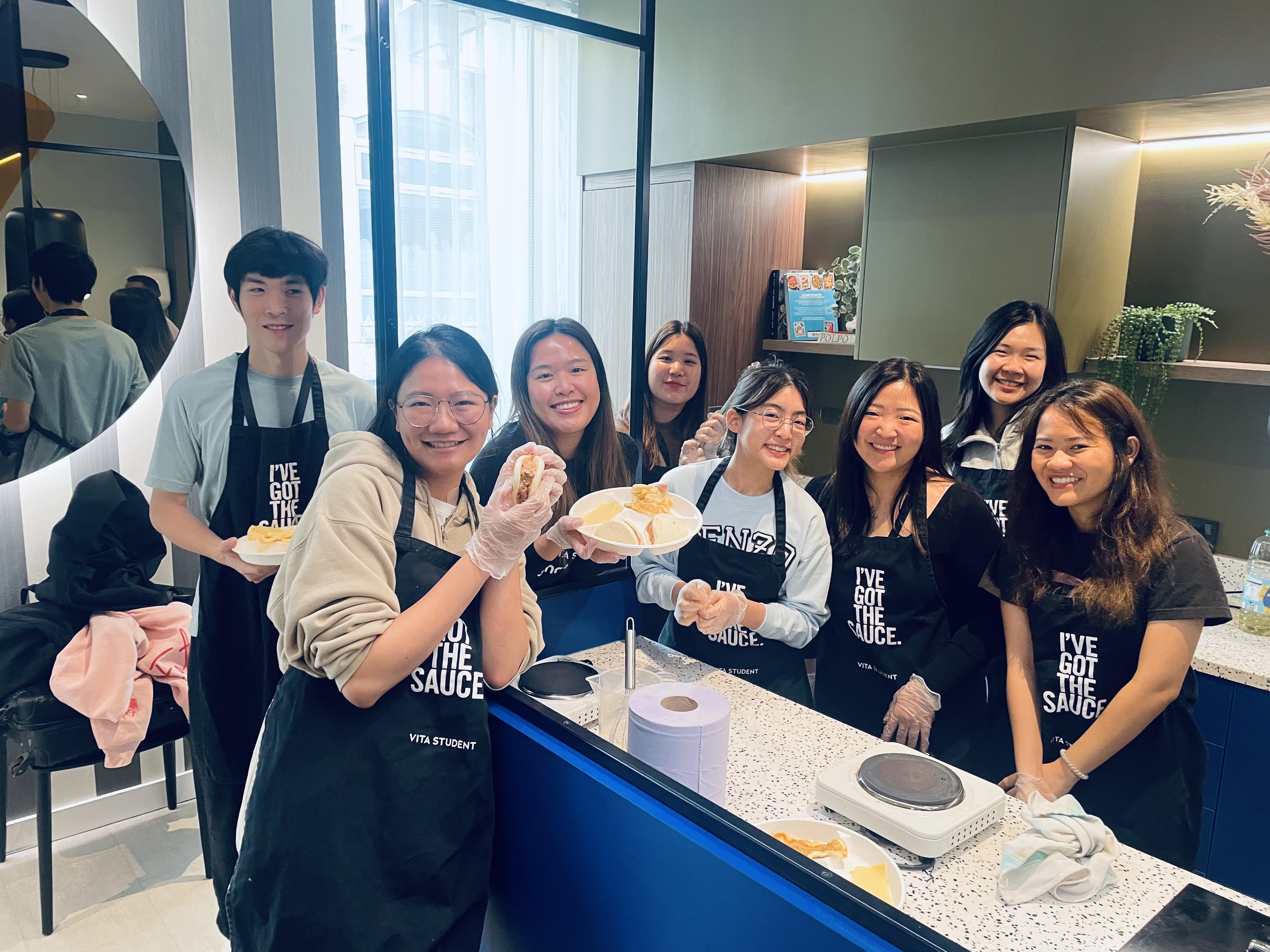 Eight smiling young people in aprons and gloves stand behind a counter at Vita Student Copper Towers, holding plates of food in a bright kitchen. Some flash peace signs, while a scale and paper towels sit on the counter—showcasing vibrant student living.