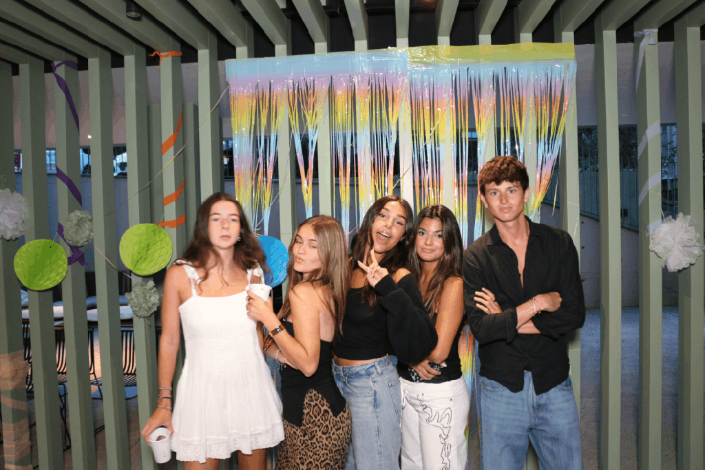 Five young adults pose in front of a colorful streamers backdrop at a Vita Student party. Four women stand together, some smiling and making peace signs, while a man in a black shirt stands on the right, celebrating the vibrant student experience.