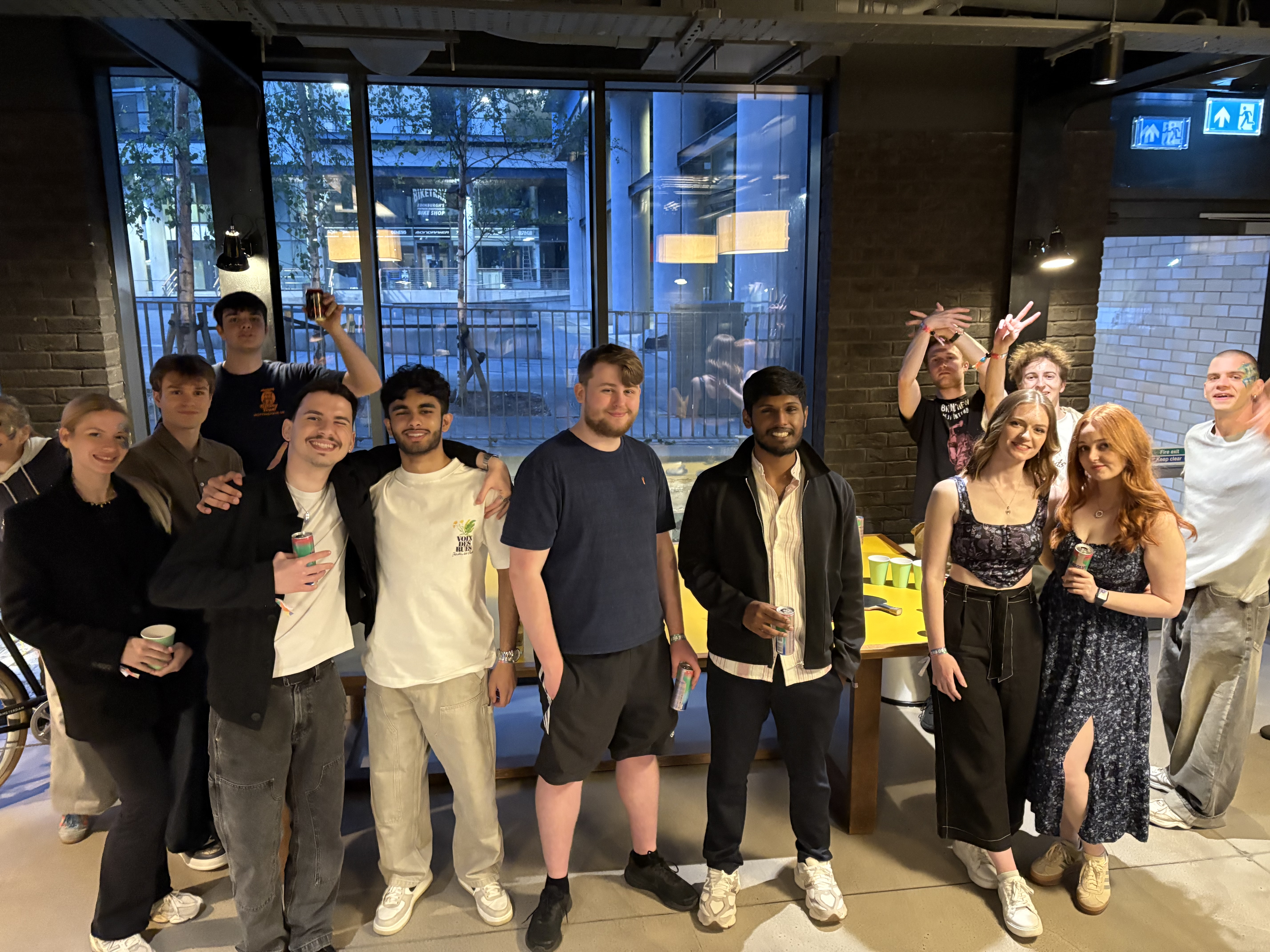 A group of young adults stands and smiles around a yellow foosball table in a modern Vita Student space with large windows. Some hold drinks, while a few in the back flash peace signs. The atmosphere looks friendly and relaxed.
