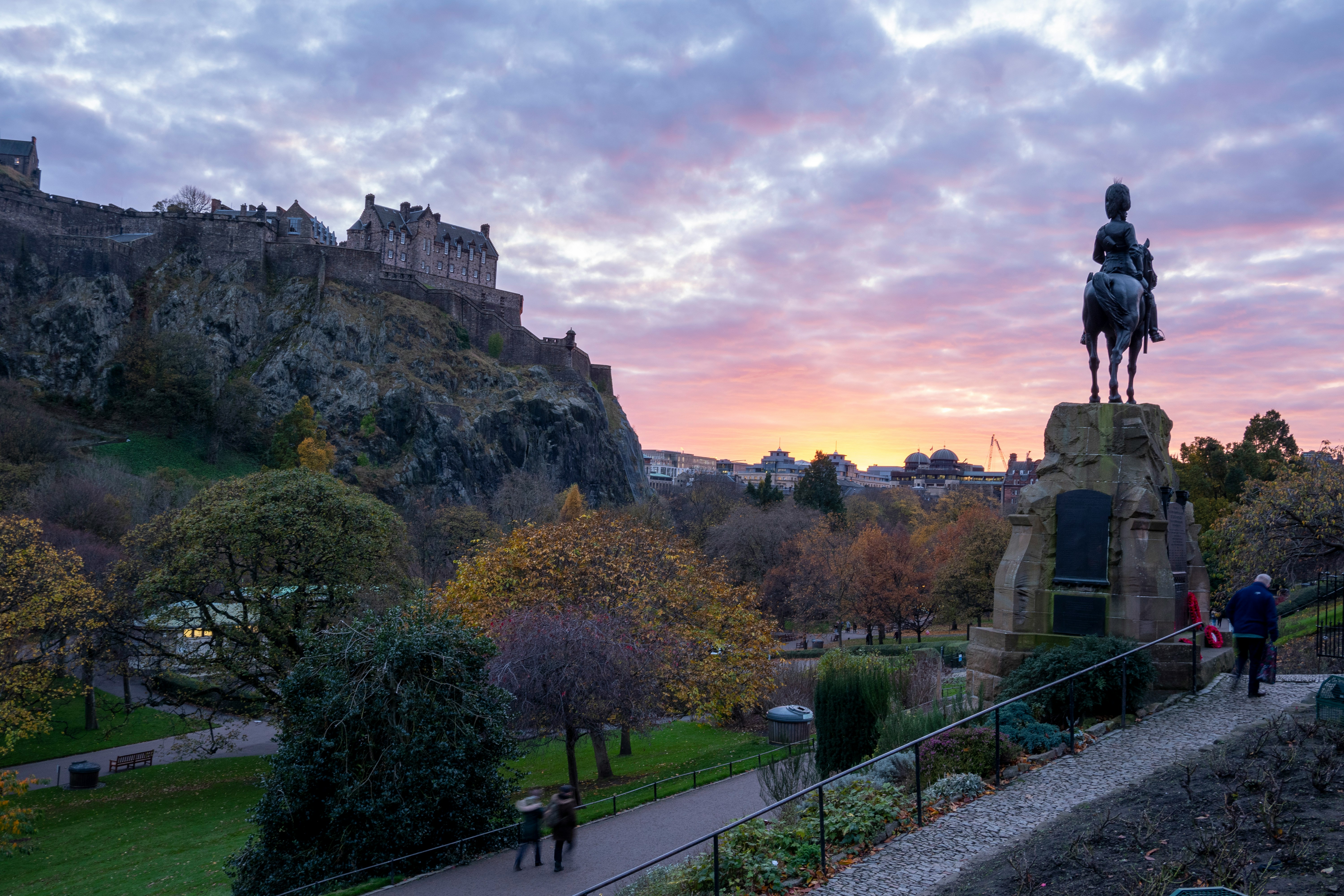 View of Edinburgh Castle on a hill at sunset, with autumn trees below, a park path curving through the foreground—perfect for students exploring things to do in the city. A statue of a rider on horseback is silhouetted against the colorful sky near New Waverley.
