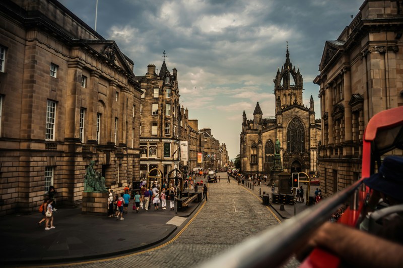 A bustling street scene in Edinburgh, Scotland, with historic stone buildings and students near New Waverley walking past St Giles’ Cathedral, under a cloudy sky.
