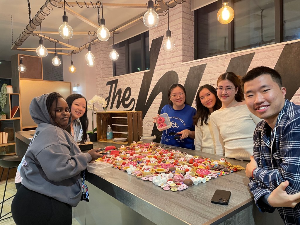 Six smiling people stand around a table covered with decorated cookies and icing supplies at Vita Student, enjoying a fun student experience in a brightly lit room with string lights and a large mural on the wall behind them.