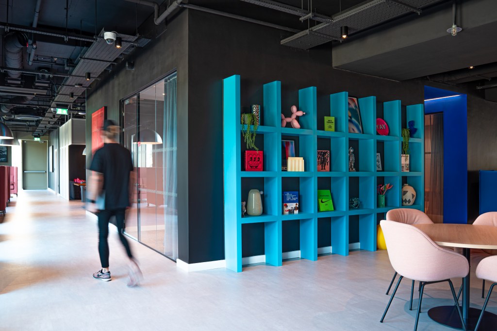 A modern office space within Vita Student Copper Towers features a blue open bookshelf filled with colorful decor, a glass-walled meeting room, pastel pink chairs around a table, and a person walking past—perfect for vibrant student living.