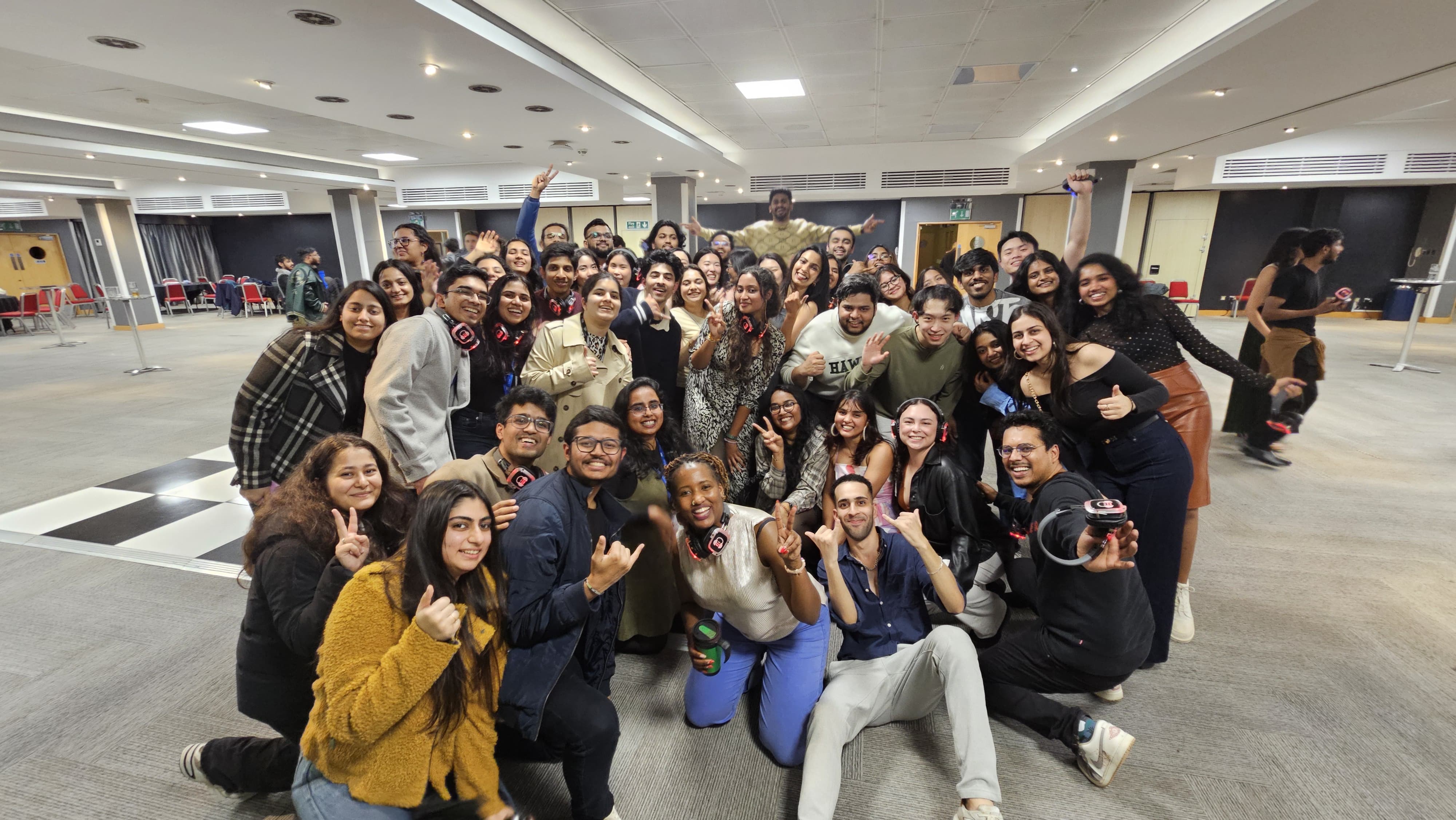 A large group of people pose together indoors, smiling and making cheerful hand gestures; the spacious, well-lit event room suggests a lively gathering where student stories are shared, with tables and chairs visible in the background.