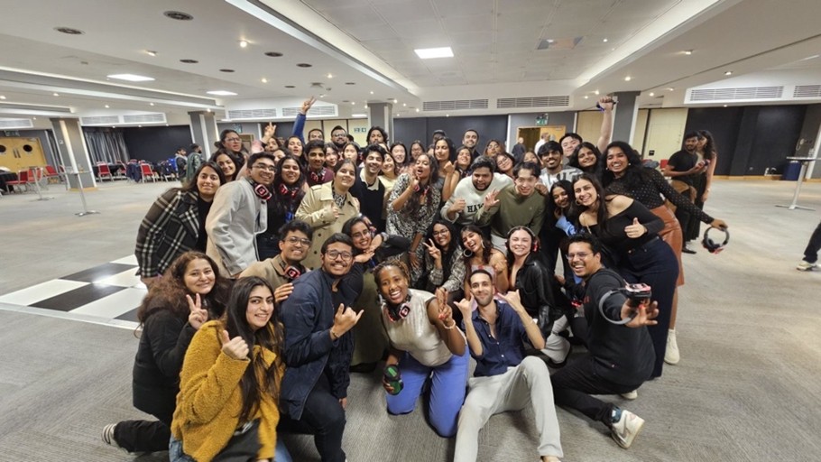 A large group of people smiling and posing for a group photo indoors, many flashing peace signs or thumbs-up gestures. Their happy faces capture the spirit of Student Stories and the leaps of faith each person has taken. A well-lit room and stacked chairs set the scene.