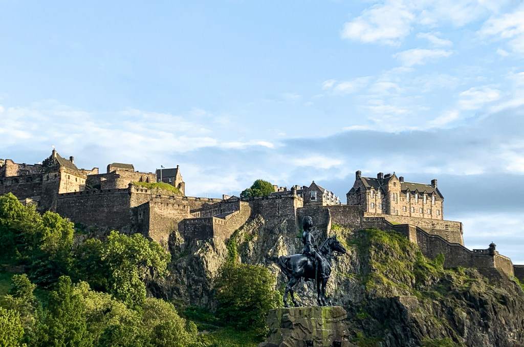 A stone castle sits atop a rocky hill, surrounded by green trees, with a statue of a person on horseback in the foreground—an iconic backdrop for student activities near New Waverley under a partly cloudy blue sky.