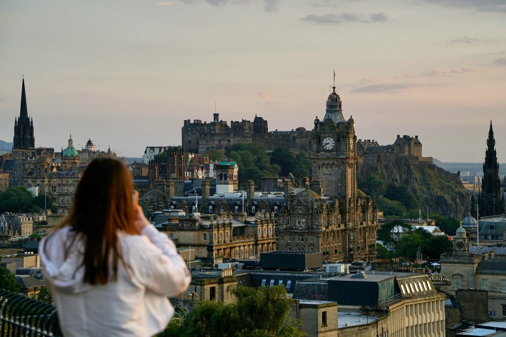 A woman in a white jacket looks at Edinburgh’s historic skyline at sunset, with the Balmoral Hotel clock tower and Edinburgh Castle beyond—one of many inspiring activities near New Waverley for students seeking stunning city views.
