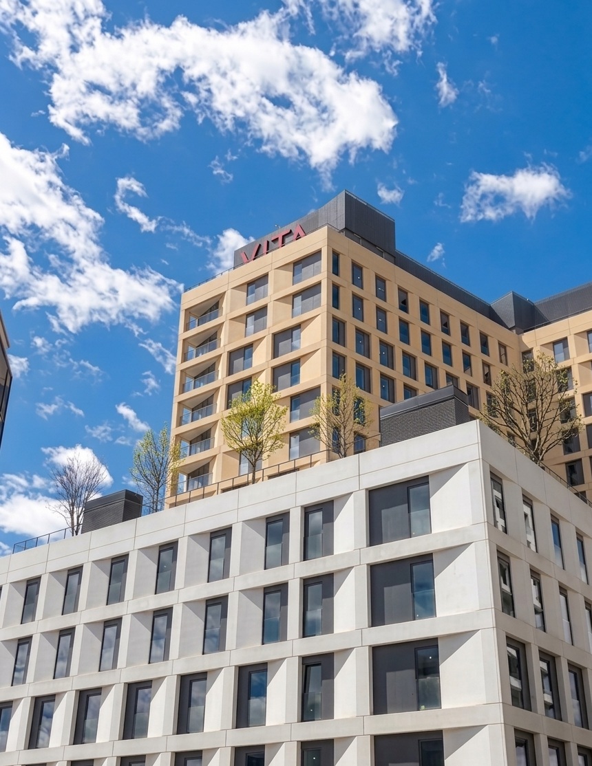 A modern multi-story building with large windows and trees on the rooftop, under a bright blue sky with scattered white clouds. The word "VITA" is visible at the top of the building.
