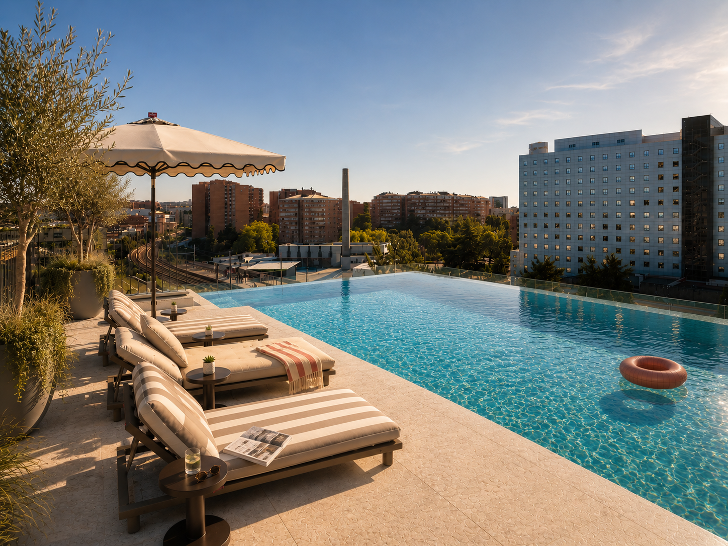 Rooftop infinity pool with striped lounge chairs, umbrellas, and a towel, overlooking a cityscape with buildings and trees; a pink pool float sits in the water under a blue sky.