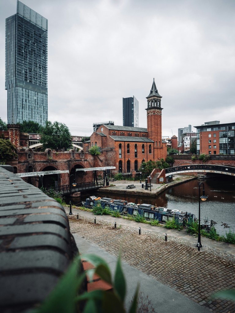 A riverside scene in Manchester features a cobblestone path, historic red-brick buildings, a modern skyscraper, canal boats, and a stone bridge—capturing the charm found near some of the best places to live in Manchester.