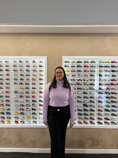 A woman in a lavender sweater and black pants stands smiling in front of two display cases filled with rows of small model cars, ready to share student stories about taking a leap of faith.