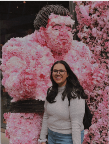 A woman with long dark hair, glasses, and a light sweater stands smiling in front of a large sumo wrestler statue made entirely of pink flowers, celebrating student stories of taking a leap of faith.