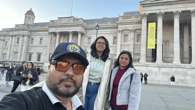 Three people pose for a photo in front of the National Gallery in London. Capturing a leap of faith, the man wears sunglasses and a cap, while two women stand close behind him, with the gallery’s columns and banners in the background.