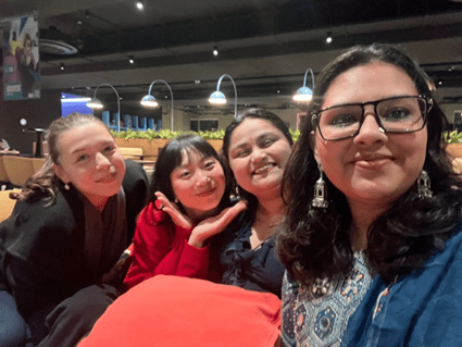 Four women smile and pose for a selfie in a well-lit indoor setting with rows of seats and hanging lamps, capturing a moment of joy that echoes inspiring student stories and shared leaps of faith.