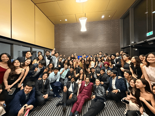 A large group of young adults dressed in formal attire pose closely together, smiling and laughing, in a modern room with brown walls and a hanging light fixture above—capturing the spirit of student stories and leaps of faith.