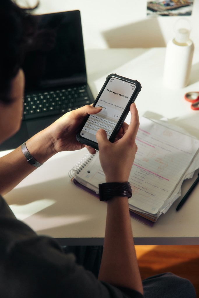 A person sits at a desk with a laptop and notebook, typing on a smartphone. The phone screen displays a message about where to live in Madrid, and there are handwritten notes and a water bottle on the desk.