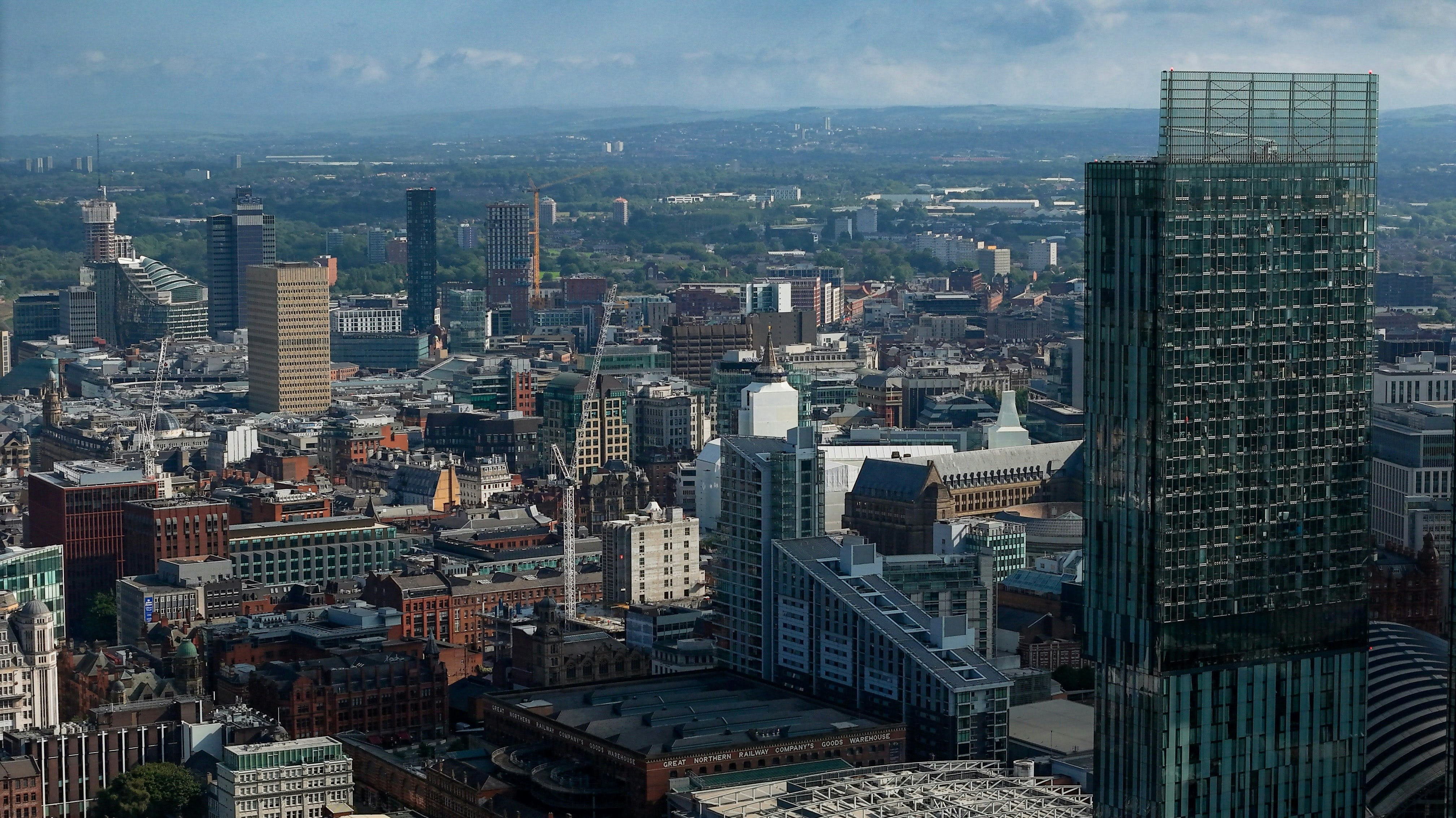 A cityscape view of Manchester, UK, featuring a tall glass skyscraper on the right, surrounded by modern and historic buildings—a familiar sight for any Manchester student—with greenery and hills visible under a partly cloudy sky.