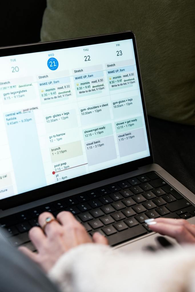 A person types on a laptop displaying a digital weekly planner with a daily schedule, including activities like gym, brunch, meetings, and researching where to live in Madrid. The planner is open to the week of the 20th to the 23rd.