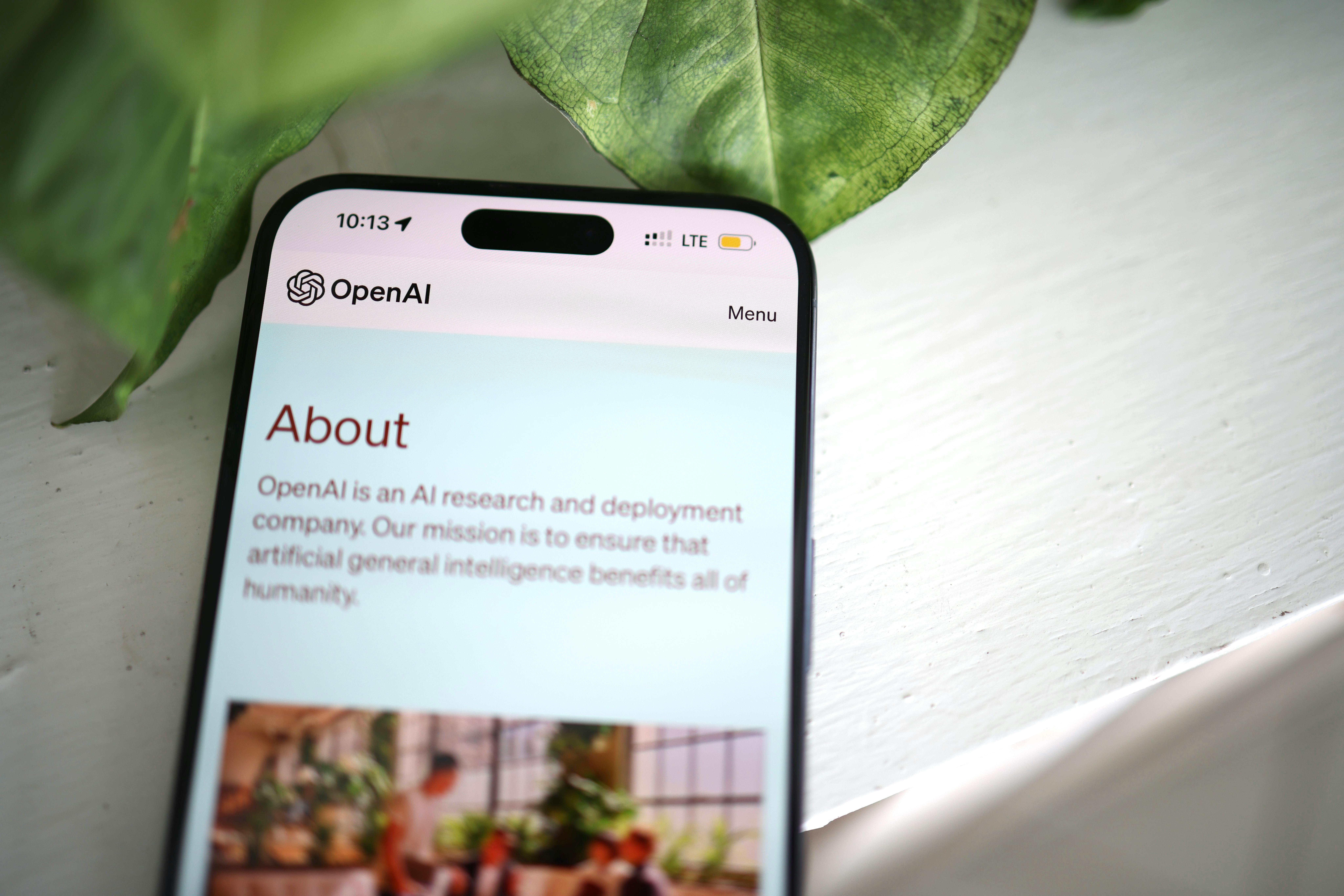 A smartphone displays the OpenAI "About" page, describing its mission to ensure artificial intelligence benefits all of humanity. The phone rests on a white surface beside green leaves, perhaps while searching for where to live in Madrid.