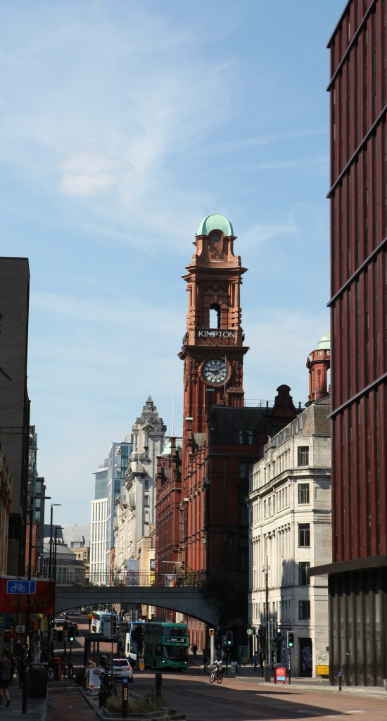 A city street with a tall red-brick clock tower labeled "KIMPTON" in the center, surrounded by historic and modern buildings—popular among students Manchester as one of the best places to live in Manchester, with buses and people on the road below.