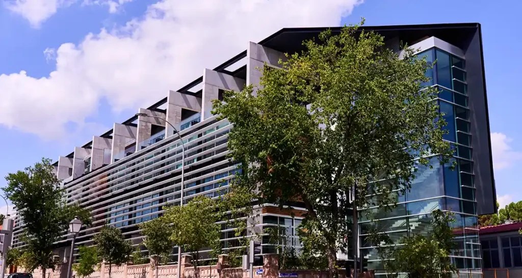 A modern glass and steel building, ideal for student accommodation in Madrid, features geometric architectural elements, large windows, and angled rooflines, partially obscured by green trees under a partly cloudy sky.