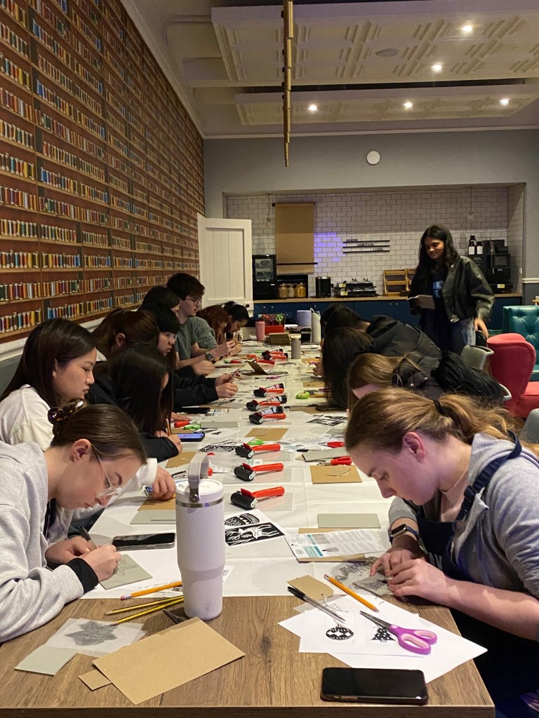 A group of people sit around a long table in York, working on arts and crafts projects with pencils and paper. The wallpaper resembles bookshelves, adding charm as one person observes—capturing genuine student stories of finding my place.