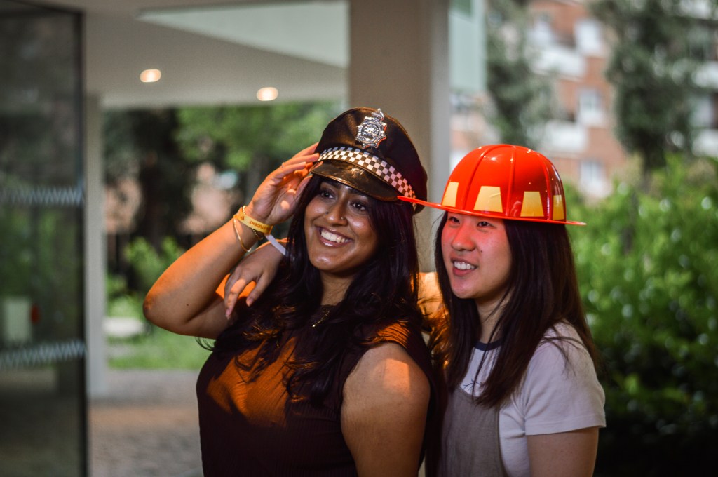 Two women smiling outdoors at Vita Student Spain, one in a police hat and the other in a red firefighter helmet. One has her arm around the other, with greenery and buildings visible in the background.