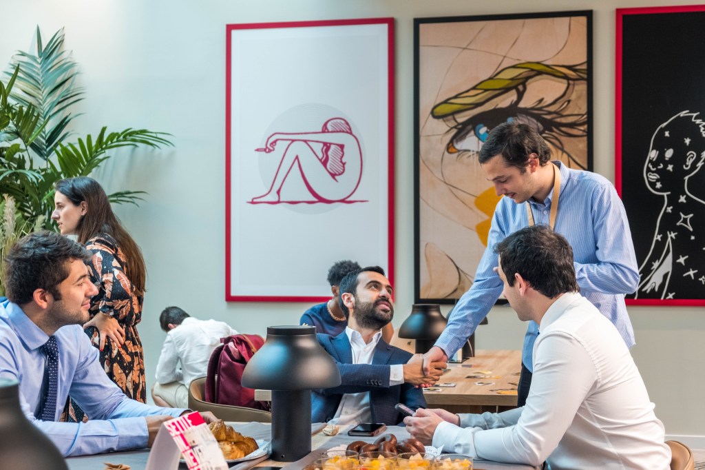 Four people, three men seated and one man standing, shake hands in a modern, brightly lit Vita Student Spain room with snacks on the table. Two large, colorful art pieces hang on the wall behind them.