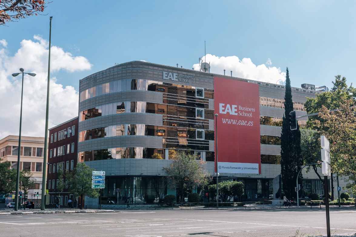 A modern glass and concrete building with a large red banner reading "EAE Business School www.eae.es" on the side, located on a city street near student accommodation in Madrid, with cars, trees, and street signs visible nearby.
