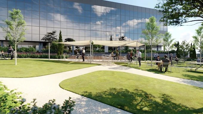 Modern office building with large reflective windows, outdoor seating under a canopy, and landscaped green lawn with winding paths. A perfect setting for student accommodation in Madrid, where people relax and chat at tables in the sunny weather.