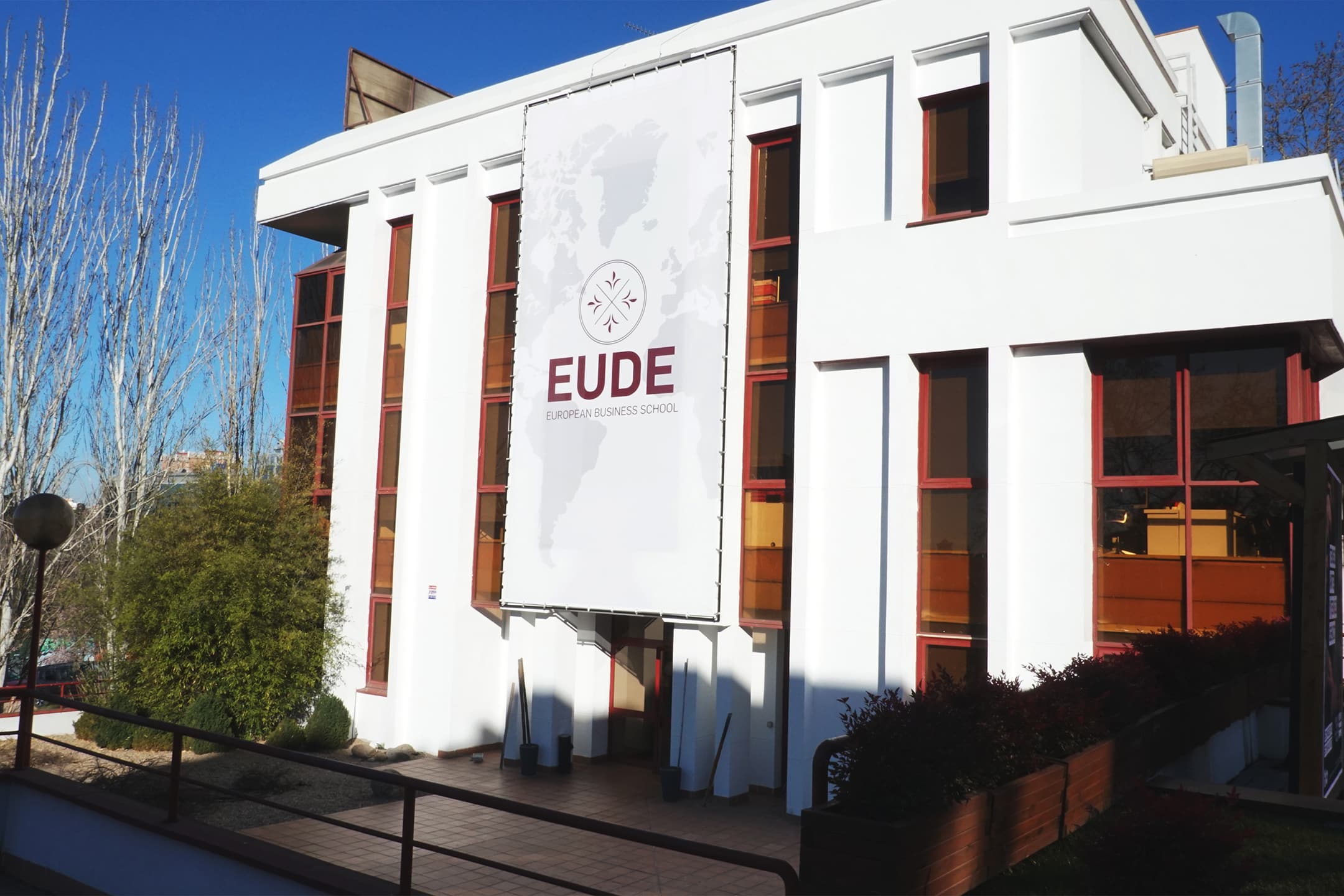 A modern white building with a large vertical banner reading "EUDE European Business School" hangs on the facade. Ideal for those seeking student accommodation in Madrid, it features tall windows and is surrounded by trees under a clear blue sky.