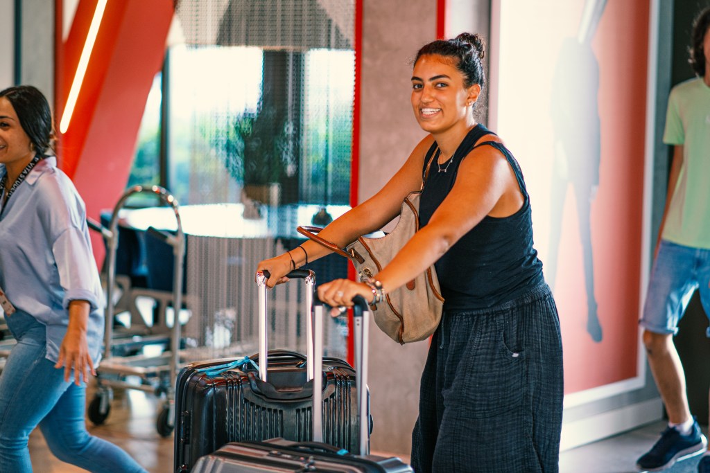 A smiling woman pushes a suitcase and carries a bag in an indoor area, possibly at Vita Student Spain or a hotel lobby. Other people with luggage are visible in the background.