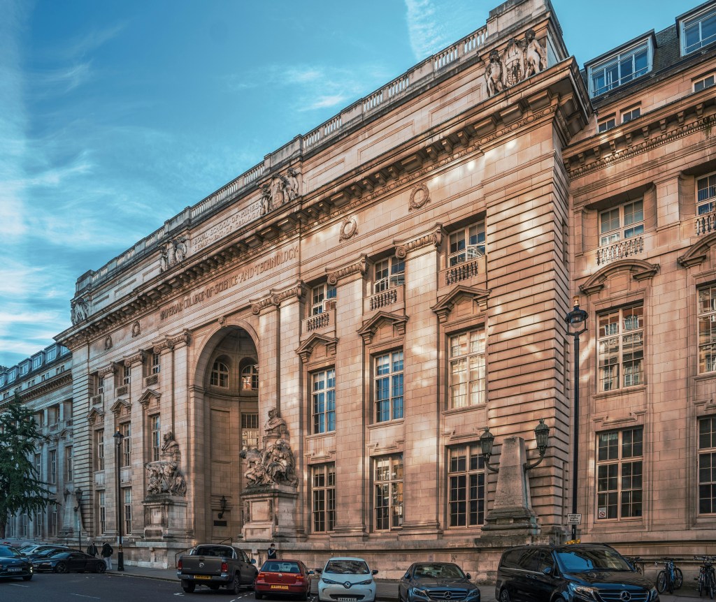 Historic beige stone building with grand columns, ornate sculptures, and arched entryway; several parked cars line the street in front under a blue sky—an iconic university featured in the QS World University Rankings 2027.