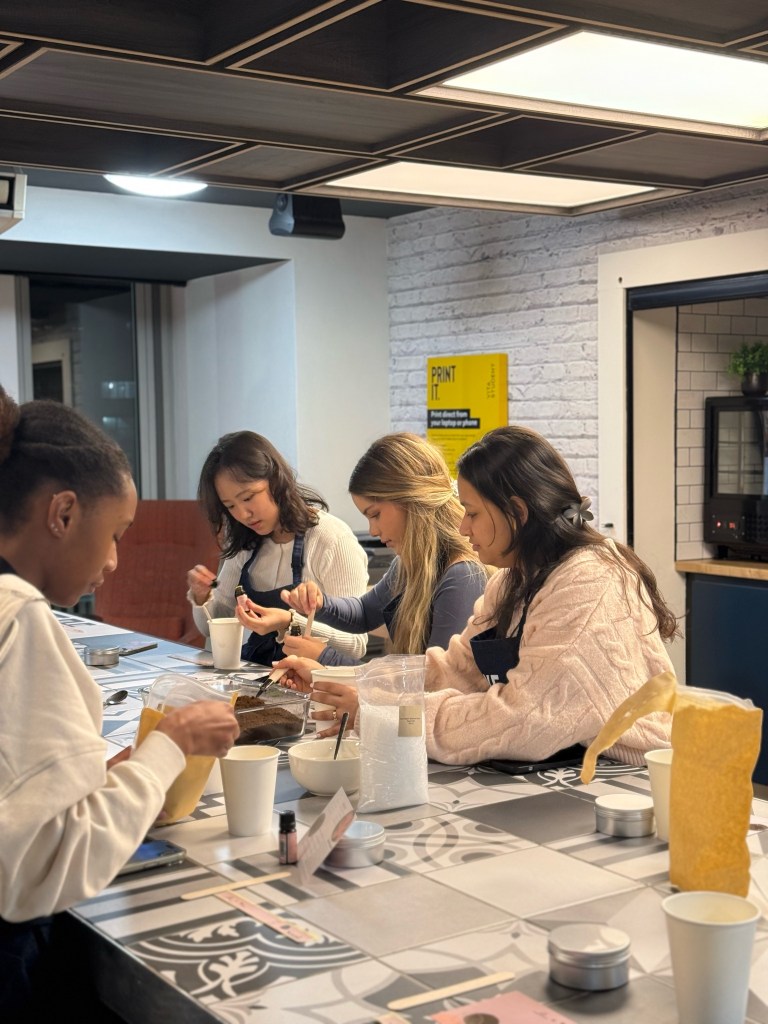 Four women sit at a kitchen counter, wearing aprons and working together on a cooking project. Their laughter and teamwork reflect how each brings a different idea of home to the modern, brightly lit setting.
