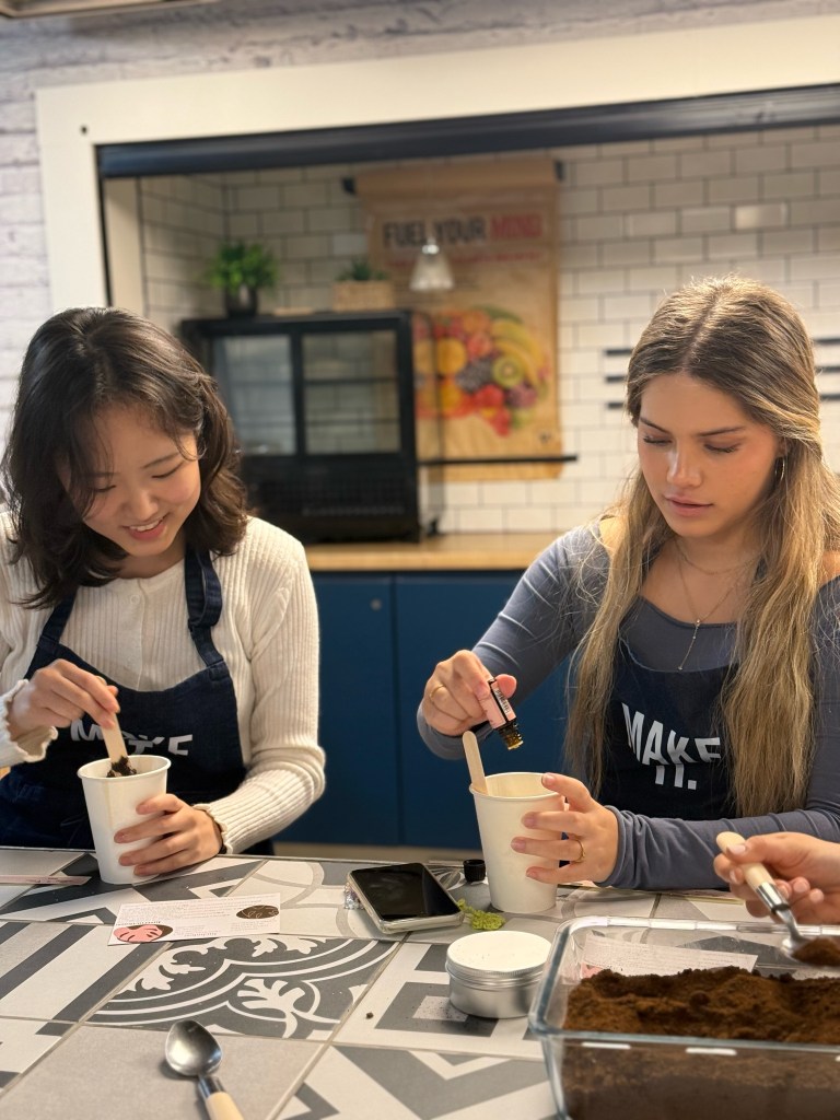 Two young women sit at a table, focused on making DIY crafts. Both are stirring cups with wooden sticks and one is adding a liquid ingredient. Their "Make It" aprons and creative supplies reflect each student's different idea of home.