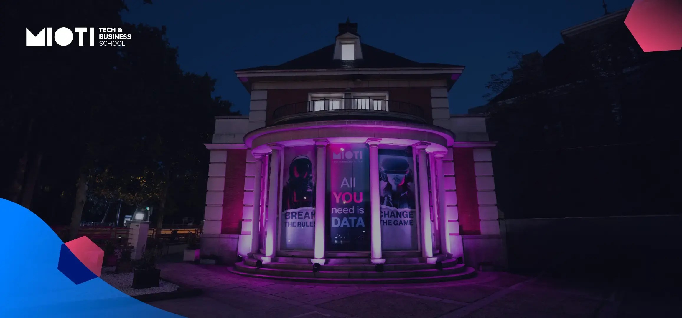 A building with columns is illuminated by purple lights at night. Large posters on the windows display the MIOTI Tech & Business School logo and slogans about data, highlighting its proximity to Student Accommodation in Madrid.