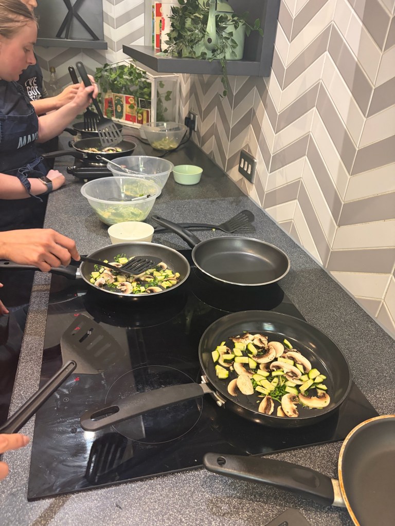 Student Stories: People cooking on a stovetop with several black pans, sautéing sliced mushrooms and zucchini. Bowls, utensils, and fresh ingredients line the counter in a modern York kitchen with patterned tile walls.