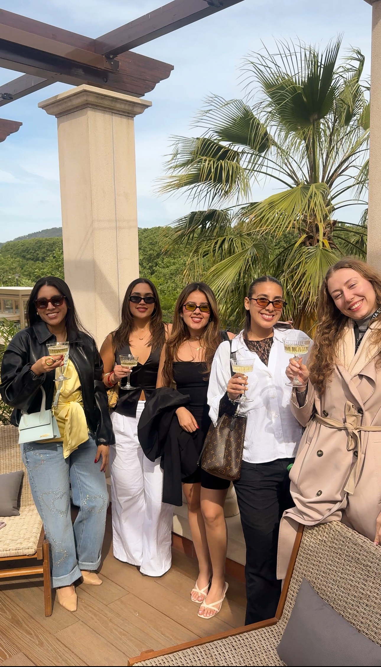 Five women stand outdoors, smiling and holding drinks during a stylish 48 hours at Vita Student Spain. They wear trendy outfits and sunglasses, gathered by palm trees and a stone column with greenery and hills behind them on a sunny day.