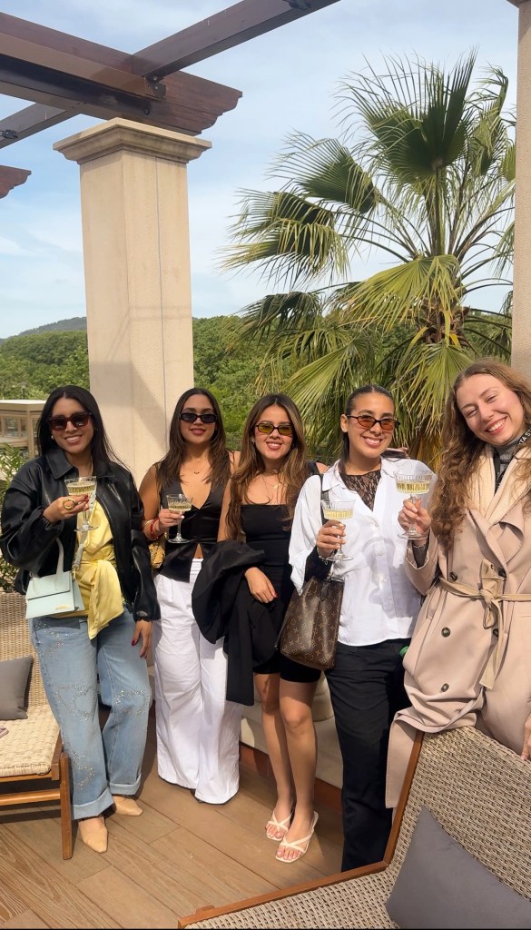 Five women stand outdoors, smiling and holding drinks during a stylish 48 hours at Vita Student Spain. They wear trendy outfits and sunglasses, gathered by palm trees and a stone column with greenery and hills behind them on a sunny day.