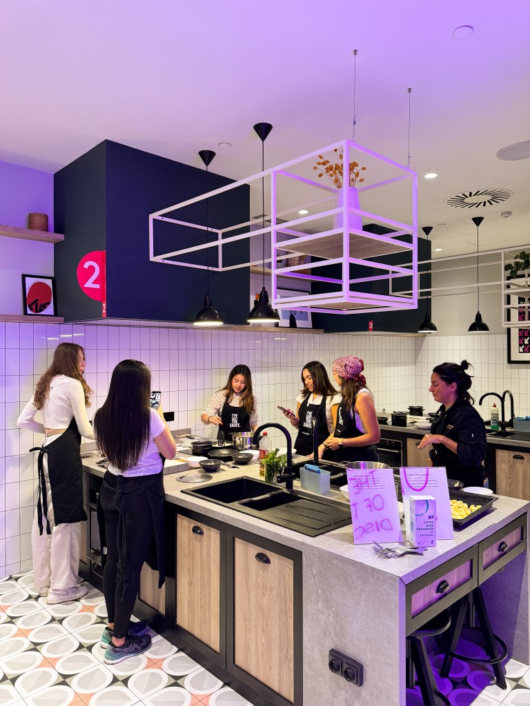Six women wearing aprons stand around a kitchen island, cooking and chatting in a modern, brightly lit Vita Student Spain kitchen—showing how student accommodation can become home in just 48 hours.