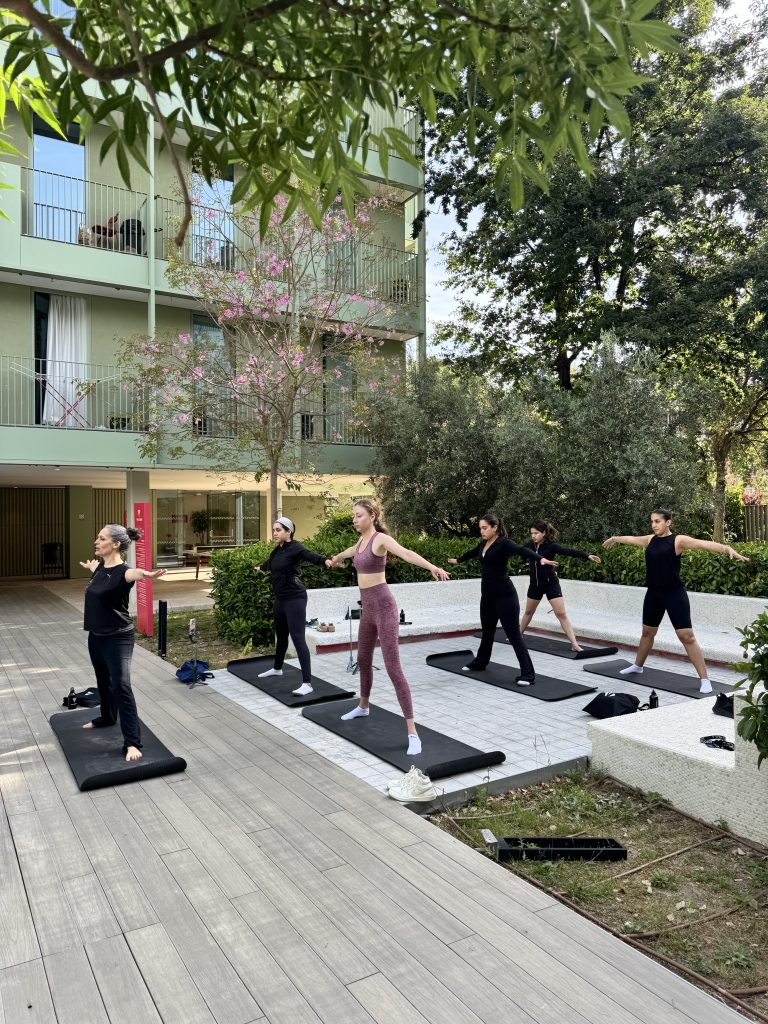 Five people practice yoga on mats outdoors near Vita Student Spain, a modern student accommodation with balconies and surrounded by trees. A yoga instructor leads the group as all participants stand in a pose with arms outstretched.