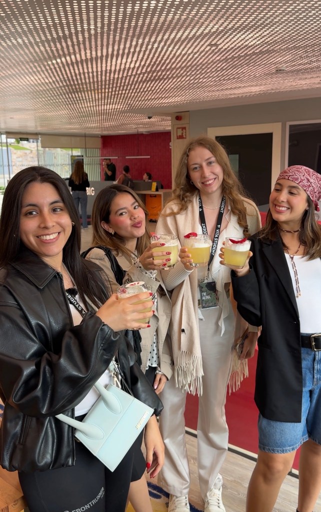Four women stand close together indoors at Vita Student Spain, smiling and holding drinks with whipped cream and strawberries. Dressed in casual, stylish outfits—one with a pink bandana—they enjoy student accommodation life in a bright, modern room.