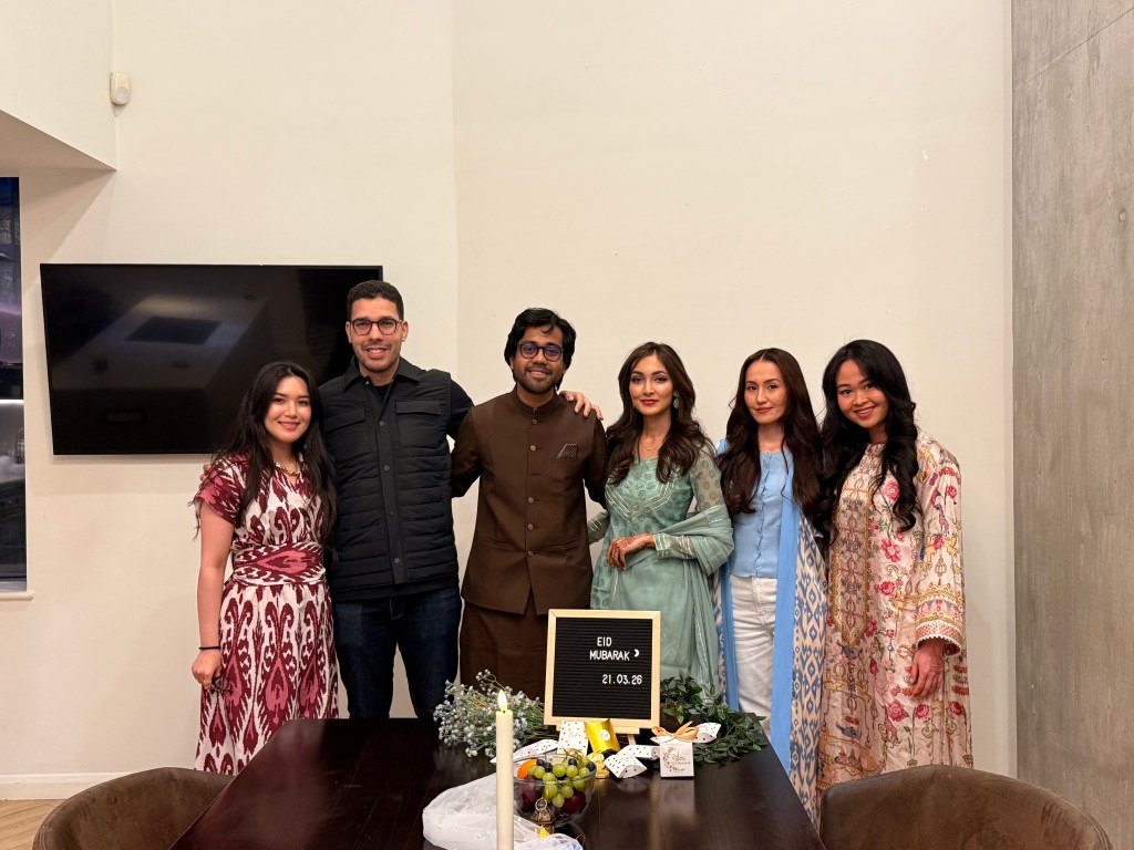 Six people stand smiling behind a dining table decorated with food and an “Eid Mubarak” sign. Dressed in festive clothing, they celebrate together, capturing joyful moments that highlight their Chevening journey in a warmly lit room.